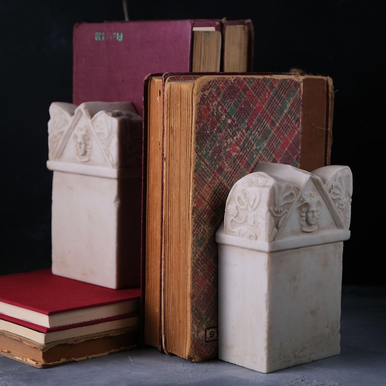 Puede incluir: Dos sujetalibros de piedra color hueso con una parte superior en front&oacute;n que presenta un relieve central de un rostro y motivos florales. Los sujetalibros sostienen una colecci&oacute;n de libros de tapa dura con cubiertas rojas y de cuadros.