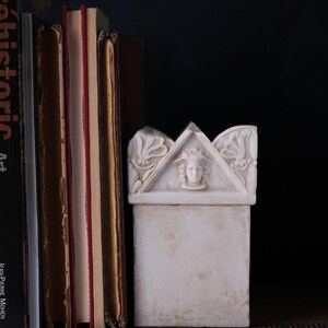Puede incluir: Una caja rectangular de m&aacute;rmol blanco con una tapa triangular que presenta un relieve tallado de un rostro con alas y motivos florales en los laterales. La caja est&aacute; colocada junto a una pila de libros sobre un fondo oscuro.