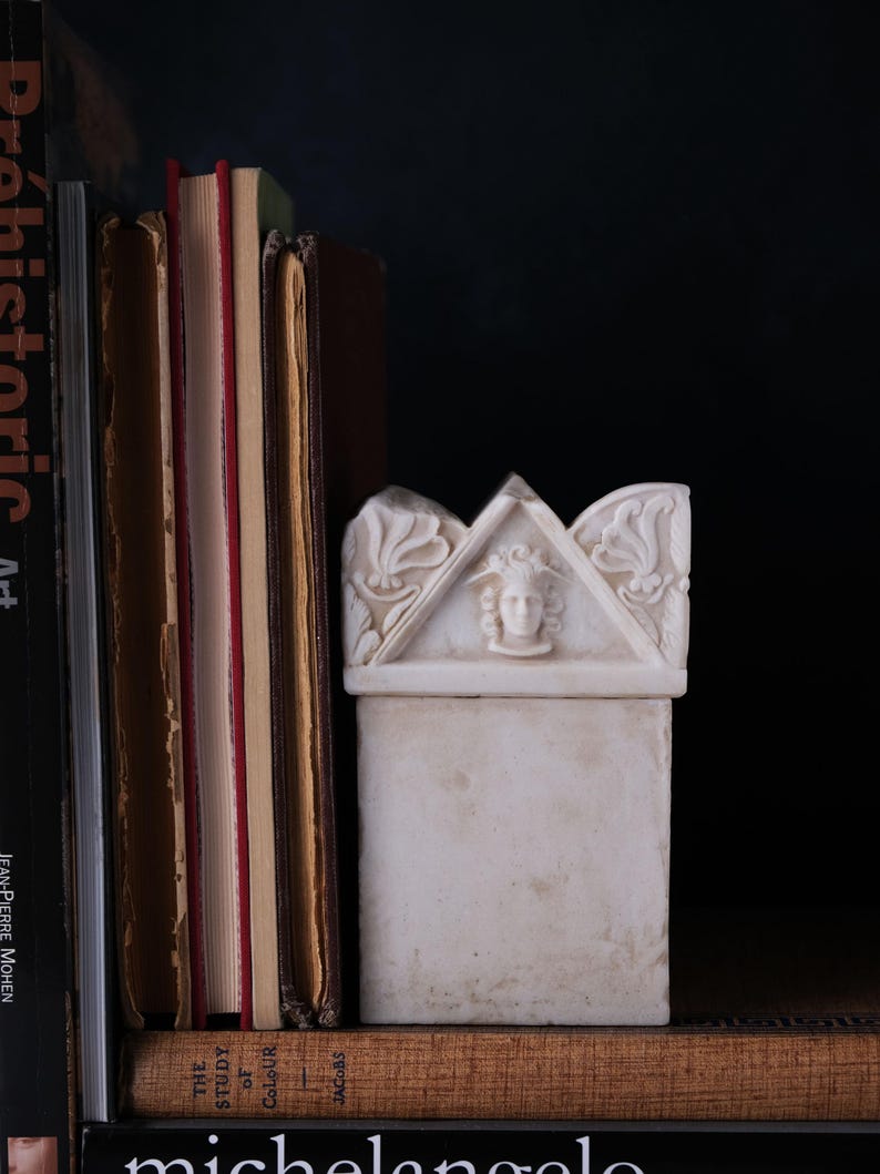 Puede incluir: Una caja rectangular de m&aacute;rmol blanco con una tapa triangular que presenta un relieve tallado de un rostro con alas y motivos florales en los laterales. La caja est&aacute; colocada junto a una pila de libros sobre un fondo oscuro.