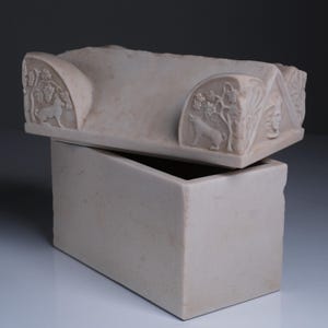 May include: A rectangular off-white stone box with a removable lid. The lid features carved relief details including a wolf standing beneath a grapevine, a human face in a triangular pediment, and a smooth, gabled top surface.
