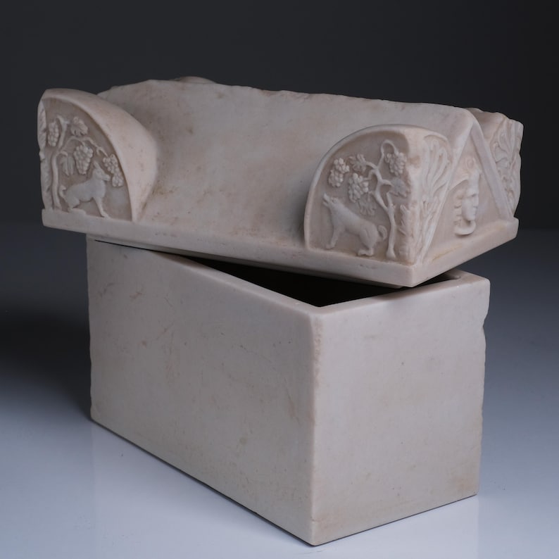 May include: A rectangular off-white stone box with a removable lid. The lid features carved relief details including a wolf standing beneath a grapevine, a human face in a triangular pediment, and a smooth, gabled top surface.