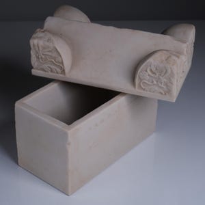 May include: A rectangular off-white stone box with a matching lid. The lid features carved floral motifs on the corners and rounded protrusions on the top edge. The box has a smooth, matte finish and a hollow interior, presented with the lid slightly ajar.
