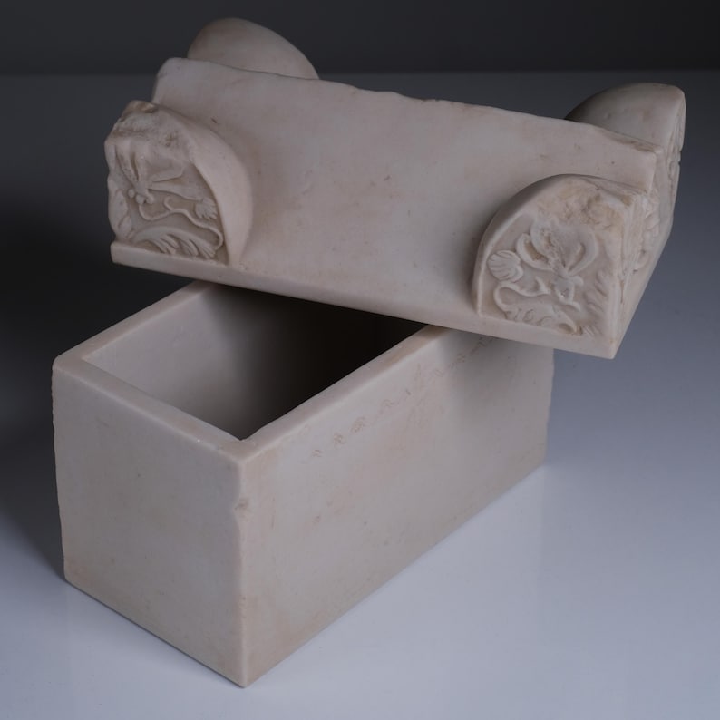 May include: A rectangular off-white stone box with a matching lid. The lid features carved floral motifs on the corners and rounded protrusions on the top edge. The box has a smooth, matte finish and a hollow interior, presented with the lid slightly ajar.