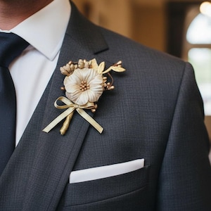 Op de afbeelding: Een antracietkleurig colbert met een wit overhemd en een donkerblauwe das. Op de revers is een corsage gespeld, bestaande uit een crèmekleurige stoffen bloem, goudkleurige metalen blaadjes, kleine gouden besjes en een dun gouden strikje.