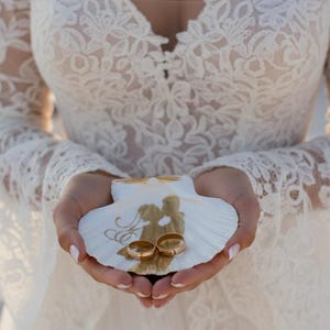 May include: Two gold wedding rings rest on a white seashell, decorated with a gold silhouette of a couple. The seashell is held in open hands, with a white lace dress in the background. The rings are a symbol of love and commitment.
