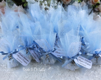 May include: A collection of light blue tulle favour bags, each adorned with a satin ribbon, a silver cross charm, and a tag. The bags are presented on a white lace surface, suitable for a baptism or wedding celebration.
