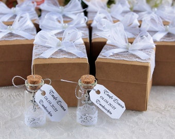 May include: Small glass bottles with cork stoppers containing silver cross charms and white lace trim, each with a tag reading Taufe Leana 04.09.2021 Dankeschön, placed in front of brown cardboard gift boxes decorated with white lace and ribbon bows.