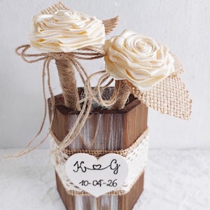 May include: A hexagonal wooden container with white distressed paint, wrapped in burlap and lace, featuring a white heart-shaped tag with the text K heart G and 10-04-26. Two cream-colored ribbon roses on stems wrapped in twine are inside.
