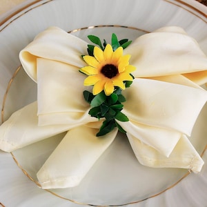 Peut inclure: Une serviette crème pliée en forme de nœud papillon, ornée d'un tournesol jaune et de feuilles vertes. La serviette est posée sur une assiette blanche avec un bord doré, pour une table élégante.