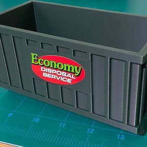 K&ouml;nnte beinhalten: Ein dunkelgr&uuml;ner Plastikcontainer mit einem roten ovalen Aufkleber, auf dem "Economy Disposal Service" in gr&uuml;nen Buchstaben steht.