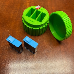 May include: A green plastic storage container with a lid, open to reveal three compartments. Two blue rectangular battery packs are sitting on a wooden surface.