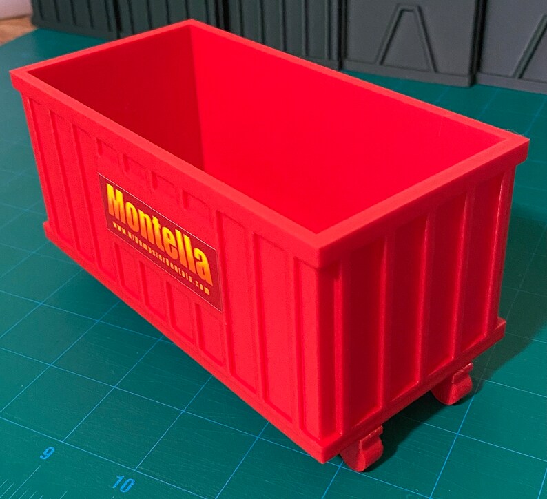 K&ouml;nnte beinhalten: Ein roter Plastikcontainer mit R&auml;dern und einem gelben und orangen Logo mit der Aufschrift "Montella" und "www.dumpstertrailers.com".