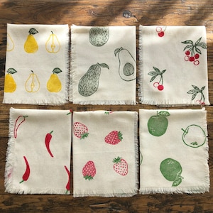 Op de afbeelding: Set van zes witte linnen servetten met verschillende fruit- en groenteontwerpen. De ontwerpen omvatten peren, avocado's, kersen, pepers, aardbeien en appels. De servetten hebben een rafelige rand.