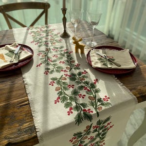 Peut inclure: Un chemin de table en lin blanc avec un motif de Noël vert et rouge représentant des branches de pin, des pommes de pin et des baies. Le chemin de table est frangé sur les bords et est posé sur une table en bois avec deux couverts.