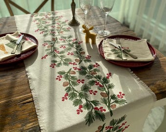 May include: A white linen table runner with a green and red Christmas-themed design featuring pine branches, pine cones, and berries. The runner is fringed on the edges and is laid on a wooden table with two place settings.
