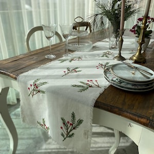 May include: A white linen table runner with a green and red floral design. The runner is on a wooden table with two wine glasses, a plate, and candlesticks.
