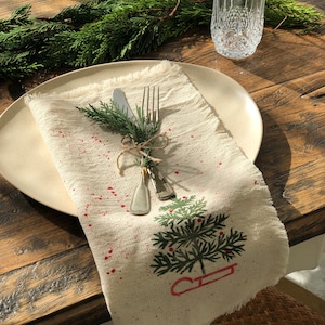 May include: A festive table setting featuring a cream-colored linen napkin with a green Christmas tree design and red splatter accents. Silverware is tied with twine and adorned with evergreen sprigs, placed on a beige plate. A glass and a decorative glass dish are also visible.
