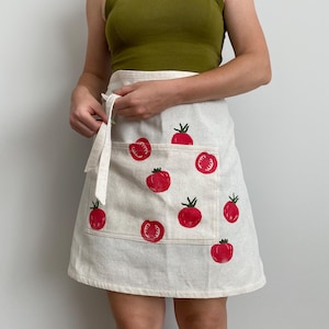 May include: A cream-coloured apron featuring a repeating pattern of red tomatoes. The apron has a pocket and a tie-around waist. The tomatoes are printed in a bold, graphic style, adding a touch of whimsy to the kitchen accessory.