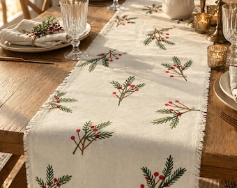 May include: A cream-colored fabric table runner featuring a repeating pattern of green pine sprigs and red berries. The runner has a frayed, fringed edge along the sides and a small rectangular brand tag attached to the bottom right corner.