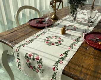 May include: A white linen table runner with a green and red floral design. The runner has a fringed edge and is laid on a wooden table with two place settings.
