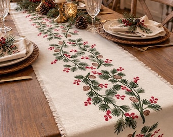 May include: A cream-colored fabric table runner featuring a printed pattern of green pine branches, red berries, and brown pinecones. The runner has a frayed edge finish and is laid out lengthwise across a wooden dining table.
