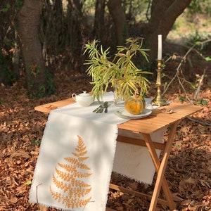 May include: A white linen table runner with a yellow fern design, placed on a wooden folding table. The table is set with a white sugar bowl, a small pumpkin, and a gold candlestick with a white candle. The table is set in a forest setting with fallen leaves on the ground.