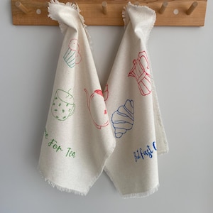 Può includere: Due asciugamani da cucina bianchi con bordo sfrangiato appesi a un portasciugamani in legno. Gli asciugamani hanno un design verde, rosso e blu con tazze da tè, teiere, pasticcini e il testo "Time for Tea" e "But first Coffee".