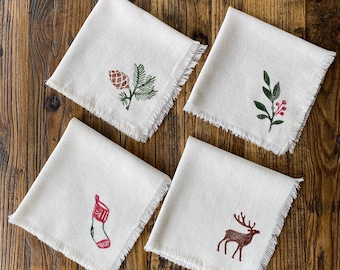 May include: Set of four white linen napkins with a frayed edge. Each napkin features a different Christmas-themed design: a pine cone and branch, a sprig of holly, a stocking, and a reindeer.
