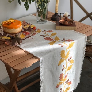 Op de afbeelding: Een witte linnen tafelrunner met een rafelige rand heeft een ontwerp van gele en oranje eikenbladeren en eikels. De runner ligt op een houten tafel met een bord oranje cake en een vaas met groen.