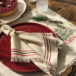 May include: A red dinner plate with a white and green striped napkin with frayed edges. The napkin is folded and held together with a gold ring. The plate is on a white placemat with a green and red embroidered design.