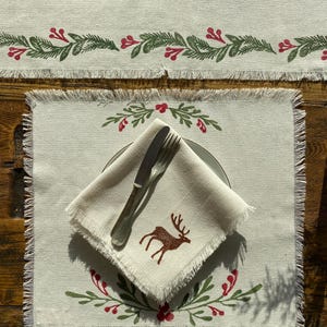 May include: Off-white linen placemat and napkin set with a rustic Christmas theme. The placemat and napkin feature a fringed edge and are decorated with green and red botanical designs and a brown deer silhouette. A fork and knife rest on the napkin.