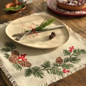 Può includere: Un piatto beige con una forchetta e un coltello legati con spago rosso, appoggiato su un sottotazza di lino con frange. Il sottotazza presenta un motivo di pigne e rami verdi e marroni, perfetto per una tavola festiva.