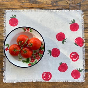 May include: A white linen placemat with a frayed edge features a red tomato design. The placemat has a bowl of red tomatoes in the upper left corner.