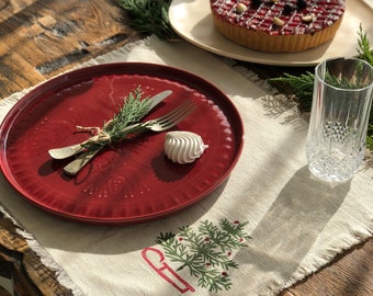 May include: A red plate with a sprig of greenery and a fork and knife on a white placemat with a green embroidered Christmas tree design. A white marshmallow sits on the plate.