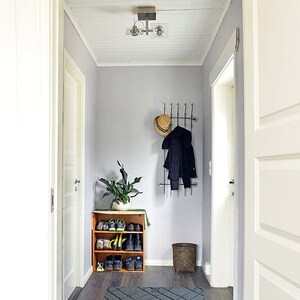 May include: A hallway with light gray walls, a dark wood floor, and white trim. A wooden shoe rack holds various shoes. A coat rack with a hat and coat hangs on the wall. A gray rug and a woven basket complete the scene.