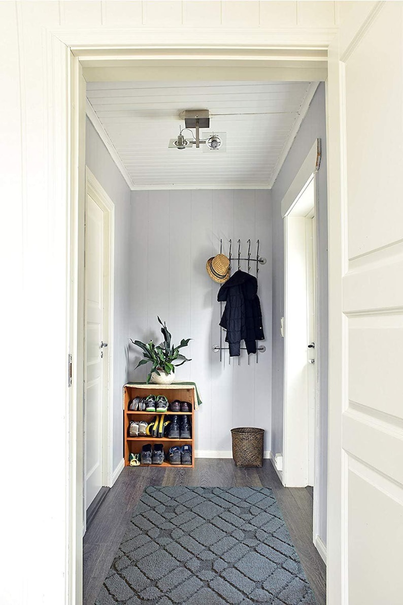 May include: A hallway with light gray walls, a dark wood floor, and white trim. A wooden shoe rack holds various shoes. A coat rack with a hat and coat hangs on the wall. A gray rug and a woven basket complete the scene.