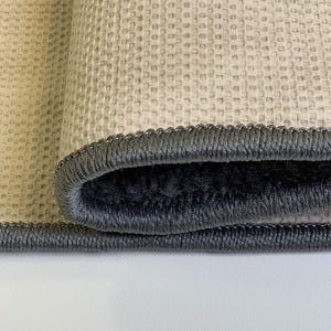 May include: Close-up of a rolled-up rug with a gray border. The rug has a textured, light beige surface and a dark gray, plush interior. The border is a darker gray with a stitched design.