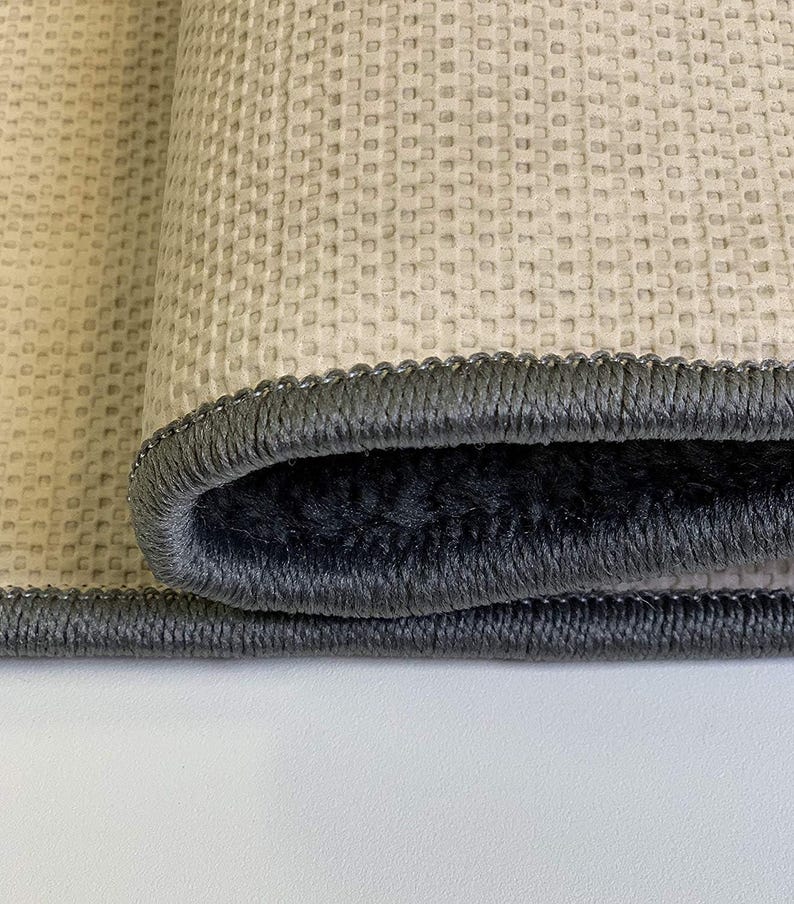 May include: Close-up of a rolled-up rug with a gray border. The rug has a textured, light beige surface and a dark gray, plush interior. The border is a darker gray with a stitched design.