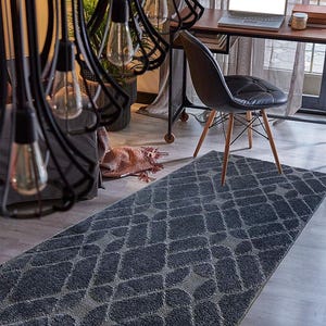 May include: A dark gray rectangular rug with a diamond pattern. The rug is placed on a light gray wooden floor. A black chair with wooden legs and a laptop on a desk are in the background.
