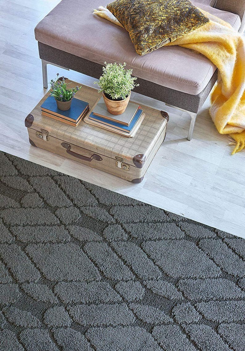 May include: A vintage suitcase serves as a coffee table, topped with books and potted plants. A gray patterned rug covers the floor, and a gray and pink bench with a yellow blanket and patterned pillow are in the background. The scene is a cozy living room.
