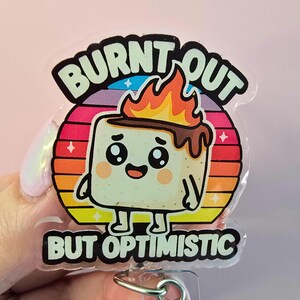 May include: A clear badge reel with a cartoon marshmallow character on a sunset background. The marshmallow has a burnt top and a smiling face. Text reads "BURNT OUT BUT OPTIMISTIC".