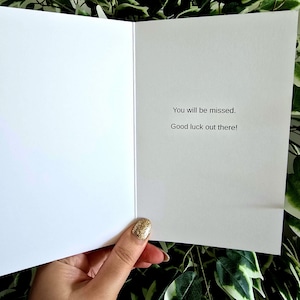 May include: An open white greeting card with the text "You will be missed. Good luck out there!" The card is held by a hand with a gold glitter nail polish. Green foliage is visible in the background.