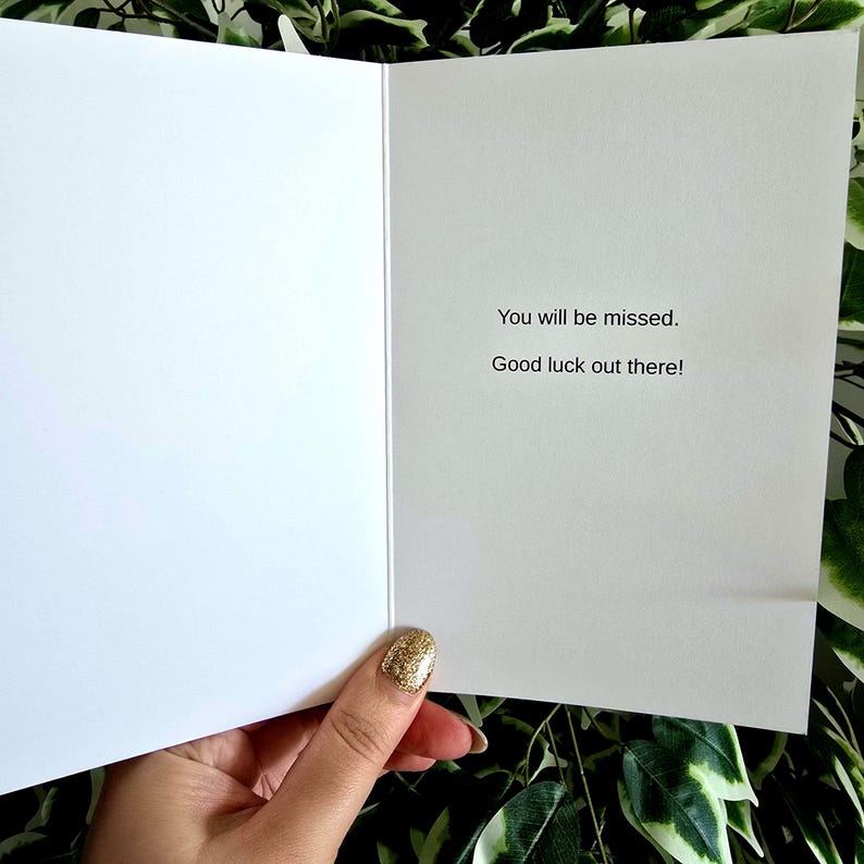May include: An open white greeting card with the text "You will be missed. Good luck out there!" The card is held by a hand with a gold glitter nail polish. Green foliage is visible in the background.