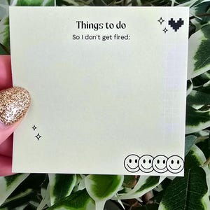 Può includere: Un blocco note quadrato, bianco sporco, con il testo "Things to do" e "So I don't get fired:" in nero. Il blocco note ha un motivo a griglia ed è decorato con simboli di cuore e stella pixelati neri e faccine sorridenti.