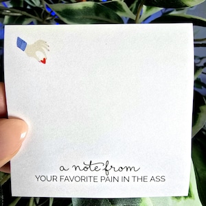 May include: A square white note card featuring a small illustration in the top left corner of a hand holding a tiny red heart. At the bottom, the text reads in black script and block letters: a note from YOUR FAVORITE PAIN IN THE ASS.