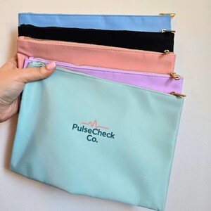 Op de afbeelding: Een stapel kleurrijke etuis met ritssluiting in lichtblauw, zwart, roze, paars en mintgroen. Op het mintgroene etui staat de tekst "PulseCheck Co.". Elke etui heeft een goudkleurige ritssluiting.