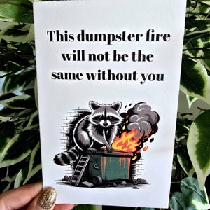 May include: A white greeting card with the text "This dumpster fire will not be the same without you." The card features a raccoon sitting on a dumpster engulfed in flames. A small ladder leans against the dumpster.