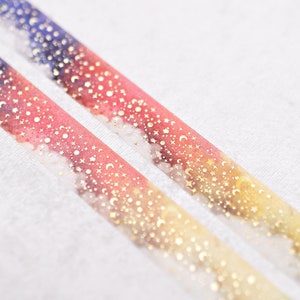May include: Two rolls of washi tape with a watercolor background of pink, orange, yellow, blue, and green. The tape is decorated with gold foil stars, moons, and dots.