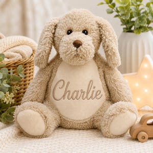 Peut inclure: Une peluche chien beige avec des oreilles tombantes, des yeux marron foncé et un nez marron foncé. Le centre de la poitrine présente un empiècement ovale beige plus clair et lisse brodé du prénom Charlie en écriture cursive marron.