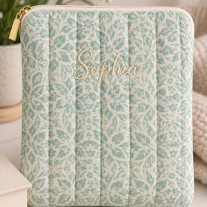 May include: A rectangular quilted pouch featuring a light green and white floral pattern. The name Sophia is embroidered in a cursive script on the front. The pouch includes a gold-toned metal zip pull and vertical stitching across the fabric.